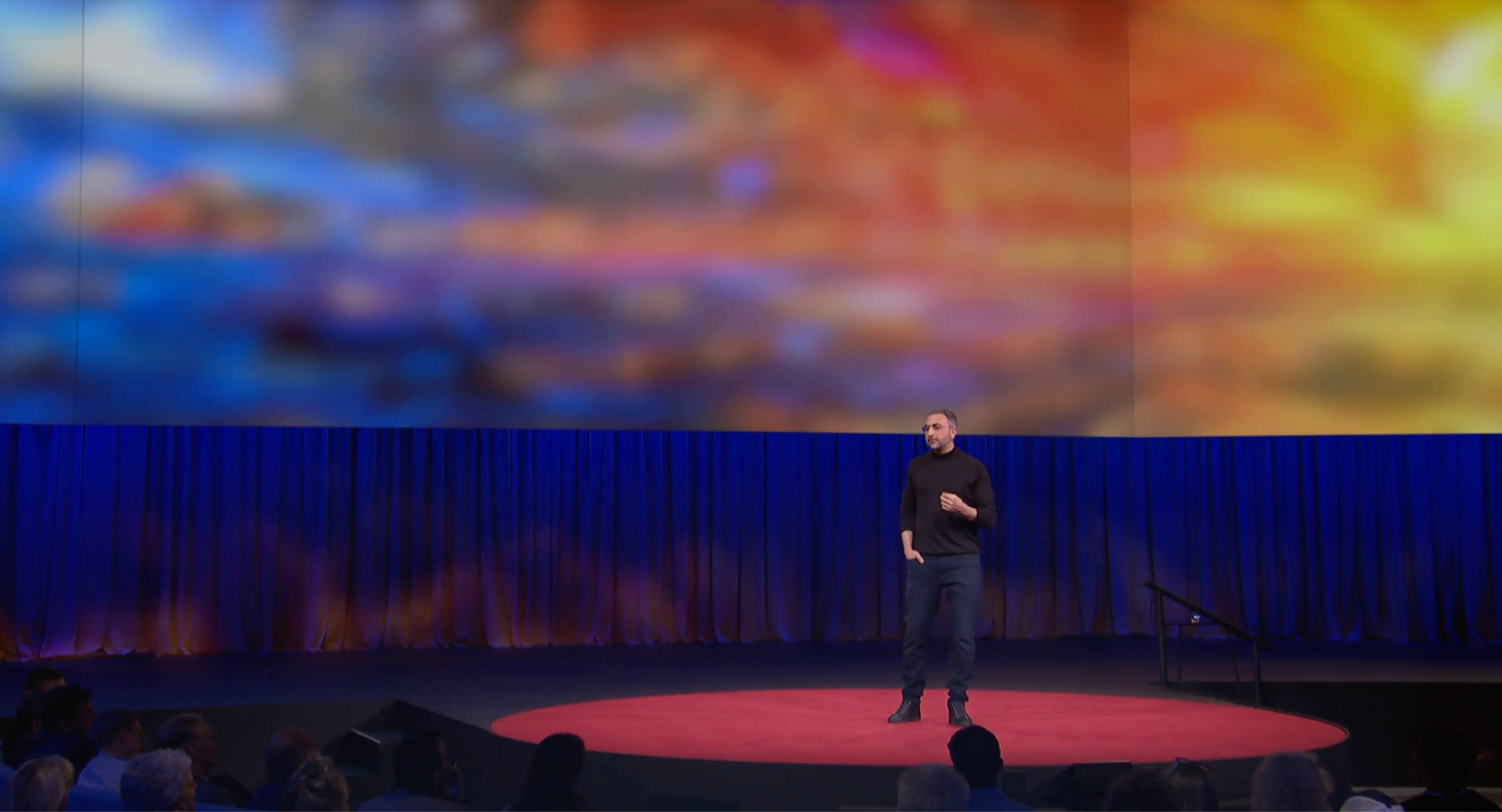 A person stands on a round red carpet delivering a talk on stage, with a large, colorful, abstract image projected behind them and an audience seated in front.